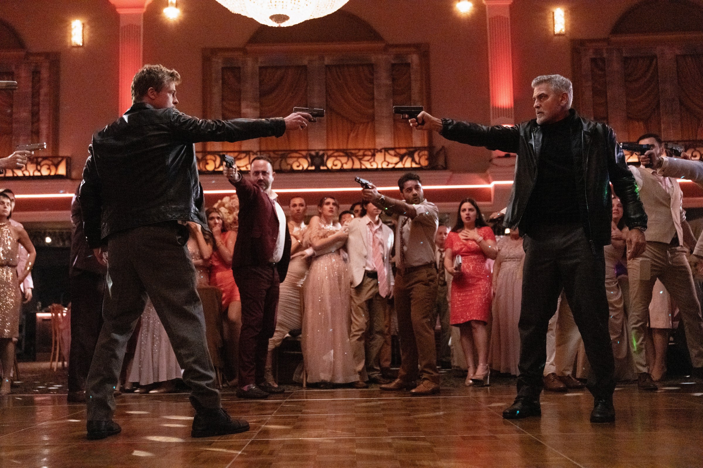 Brad Pitt and George Clooney pointing pistols at one another while other people point guns at them on a dance floor in Wolfs.