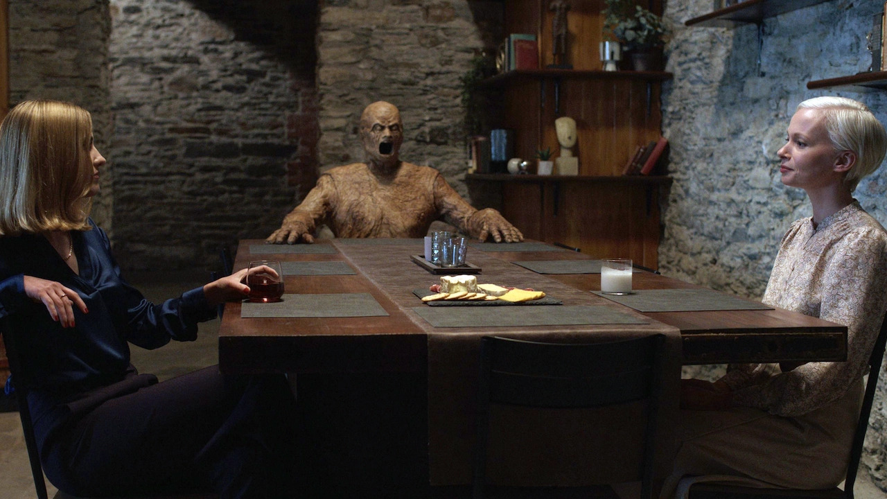 Two woman sitting across from one another at a table with a ghoulish wooden figure in Oddity.
