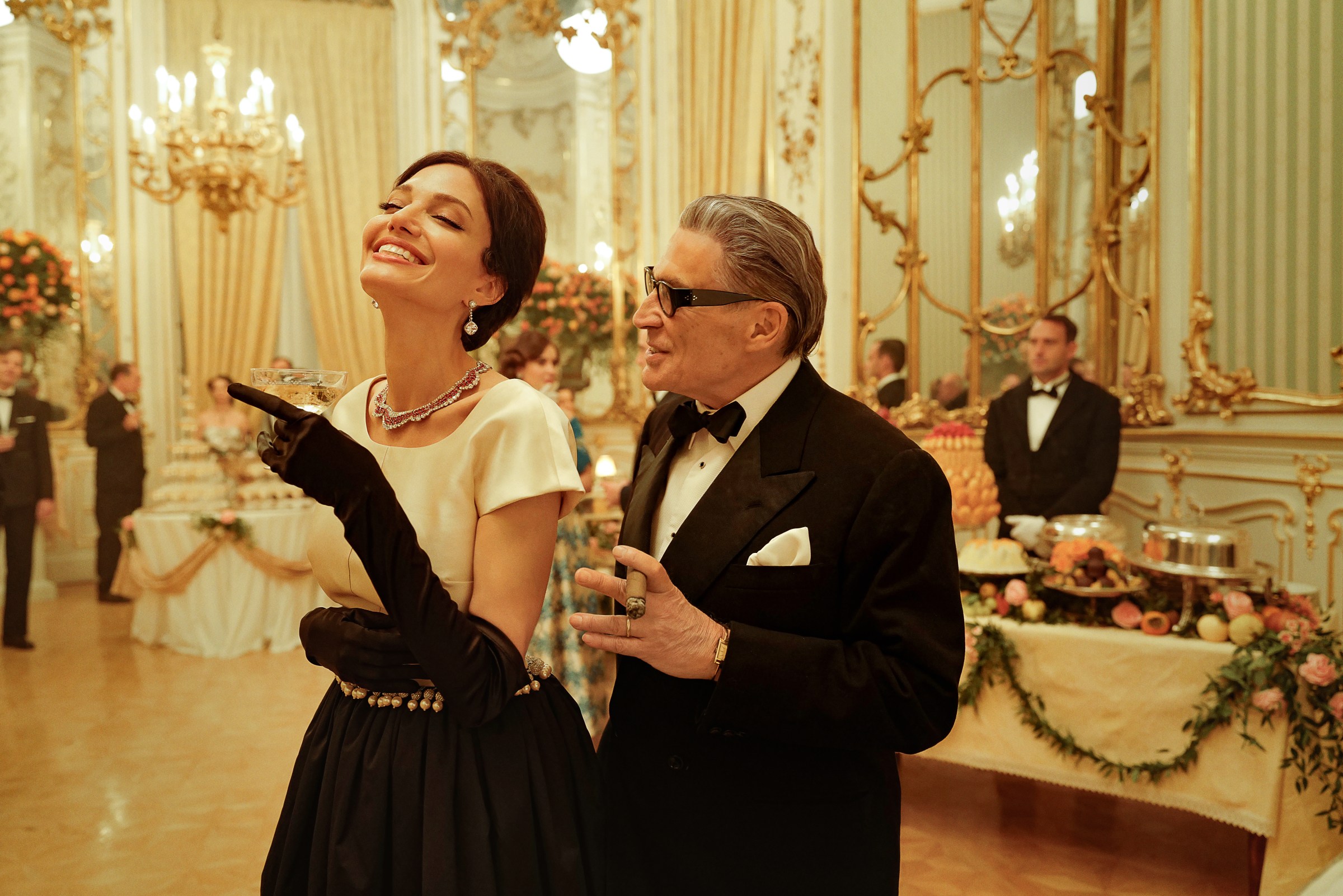 Angelina Jolie holding a champagne glass in a lavish party hall next to a man in tuxedo in Maria.